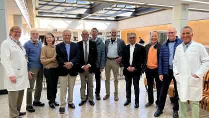 A group of people standing together indoors in a bright, modern space with large windows and skylights. Two individuals are wearing white lab coats, while others are dressed in business or casual attire. 