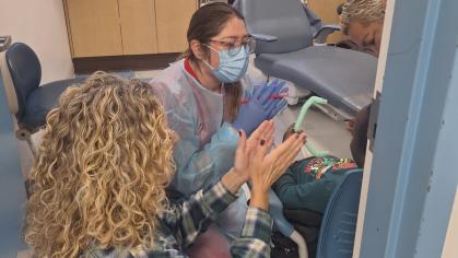 Dental clinic scene showing a masked dental professional and an adult caregiver using hand gestures to communicate with a child seated in a dental chair, while another clinician observes, with dental equipment and cabinetry visible in the background.