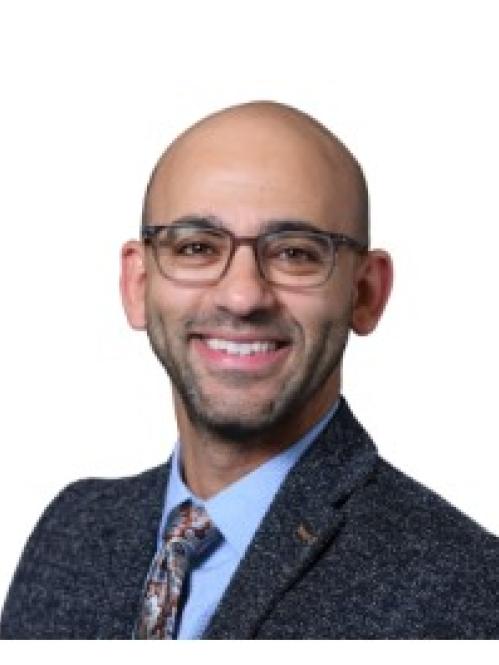 A smiling man with glasses, a bald head, and a neatly trimmed beard. He is wearing a dark textured suit jacket, a light blue shirt, and a patterned tie, posed against a plain white background.