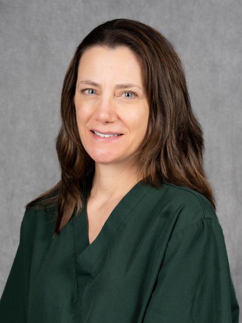 A woman with fair skin and long, straight brown hair parted slightly to the side. She is wearing a dark green scrub top and is smiling gently while facing the camera. The background is a plain gray backdrop.