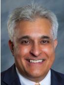 Smiling man with short gray hair wearing a dark suit, white shirt, and yellow tie, posed in front of a blurred background