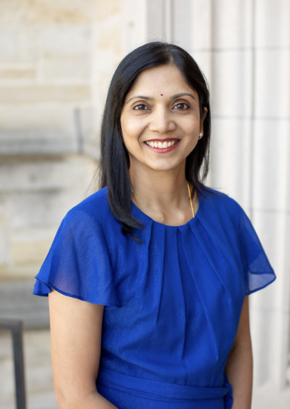 A person with long black hair is wearing a blue blouse and standing in front of a stone wall. 