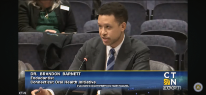 Dr. Brandon Barnett, an endodontist, speaks at a public hearing for the Connecticut Oral Health Initiative. He is seated at a table with a microphone, wearing a suit and tie.