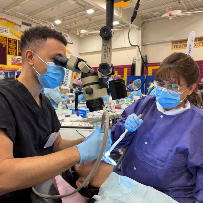 Two dental professionals perform a dental procedure in a large indoor venue. One, wearing black scrubs and a blue face mask, while the other, in a purple gown and mask, assists with instruments. 