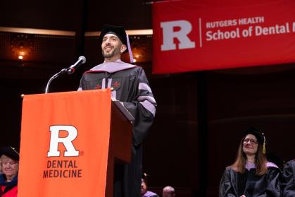 A graduate in academic regalia stands at a podium with a microphone, speaking during a ceremony. The podium displays a large red banner reading “R Dental Medicine,” and a red backdrop behind shows “Rutgers Health School of Dental Medicine,” while seated faculty members in caps and gowns are visible in the background.