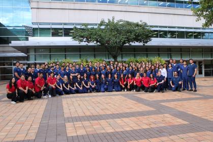 Large group photo of approximately 100 people gathered outdoors in front of a modern glass-and-metal building. Most of the group is wearing navy blue medical-style scrubs, while a smaller group in the front row is wearing red polo shirts. The group is arranged in several rows on a brick plaza beneath a large tree centered in front of the building entrance. Daylight conditions with the building, windows, landscaping, and paved courtyard visible in the background.